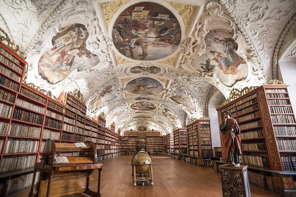 Inside the Strahov Library there is a white, ornate ceiling with religious paintings and below it are bookshelves.