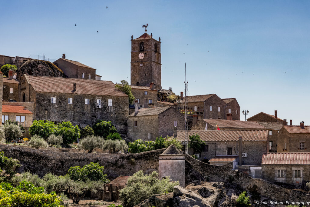 View of the main square in Monsanto Village. Birds are flying about the church tower while the sun shines in a blue sky.